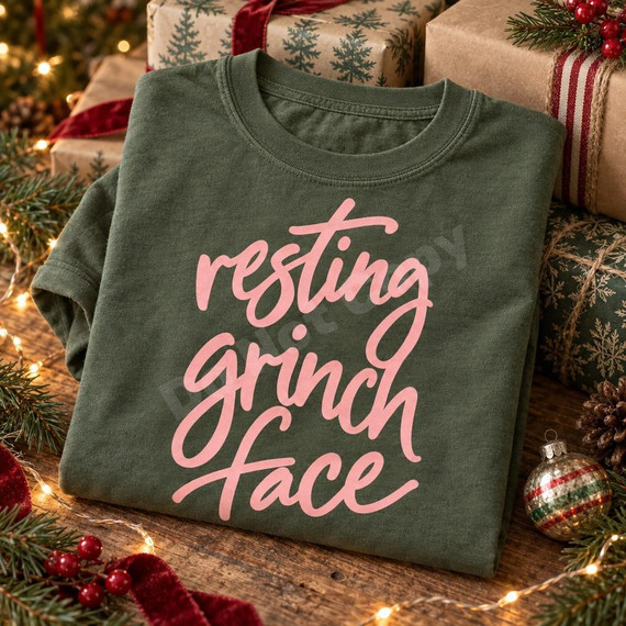 Resting Grinch Face DTF Transfer | Funny Hand-Lettered Christmas Design, pink hand-lettered “Resting Grinch Face” design displayed on a folded green shirt surrounded by festive Christmas gifts and decorations.