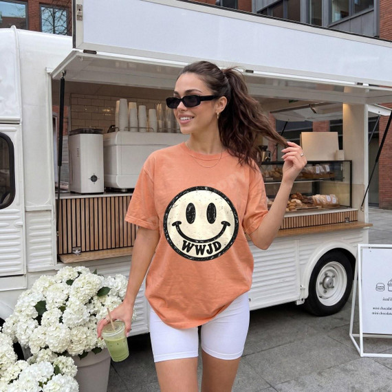 WWJD Smiley Face DTF Transfer | Retro Faith-Based Distressed Design, model wearing an orange T-shirt featuring a distressed black-and-cream smiley face with WWJD lettering