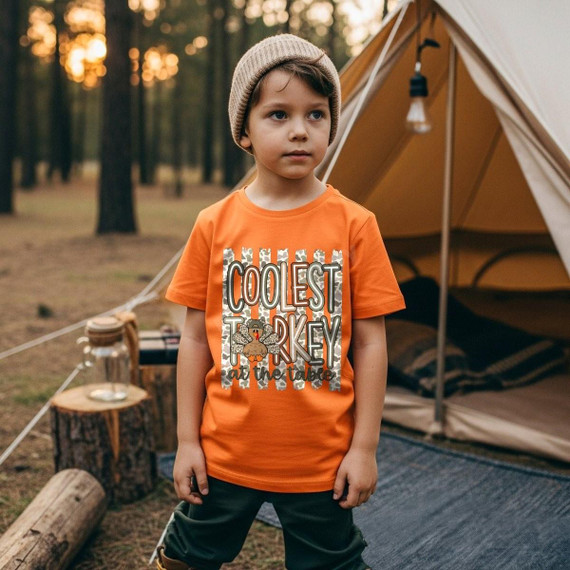 Coolest Turkey at the Table DTF Transfer | Camo Kids Thanksgiving Design, orange children’s shirt featuring a camouflage turkey and “Coolest Turkey at the Table” Thanksgiving graphic, modeled by a child at a campsite.