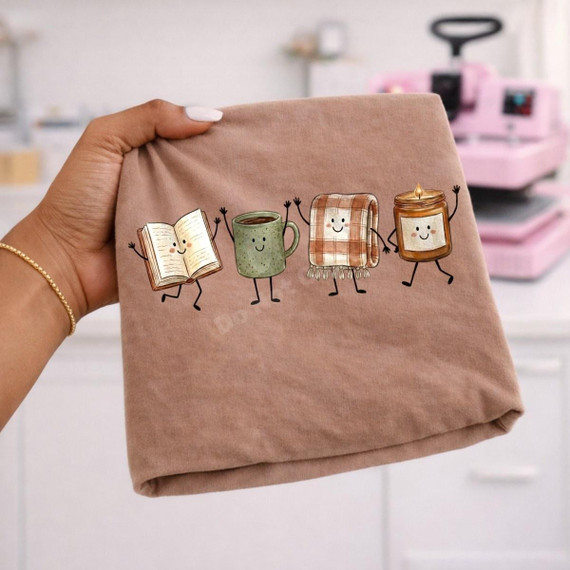 Cozy Fall Things DTF Transfer | Dancing Book, Coffee, Blanket & Candle Design, cheerful fall-themed DTF transfer featuring dancing cartoon book, green coffee mug, plaid blanket, and candle printed on a folded tan shirt.