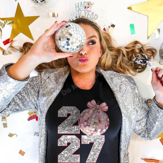 2027 Disco Ball DTF Transfer | New Year's Eve Party Design, model wearing a black shirt with a glittery silver 2027 and pink disco ball graphic while posing with a mirrored disco ball amid festive New Year's Eve decorations.