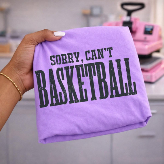 Sorry, Can't Basketball DTF Transfer | Funny Basketball Game Day Design, black distressed “Sorry, Can’t Basketball” lettering displayed on a folded purple shirt beside a heat press.