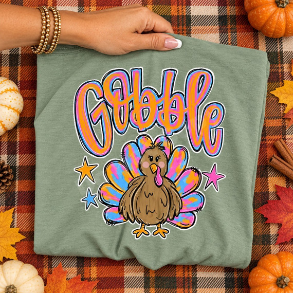 Gobble DTF transfer with cute brown turkey, tie-dye style pink, orange and blue tail feathers, bubbly lettering, and pink and orange stars Shown on a Sage t-shirt.