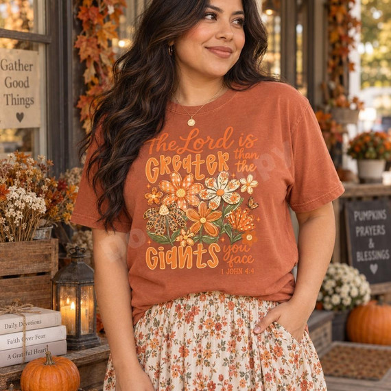 The Lord is Greater Than the Giants You Face DTF Transfer | Faith Floral Design, faith-inspired floral graphic displayed on a rust-colored T-shirt worn by a woman, with the Bible verse 1 John 4:4.