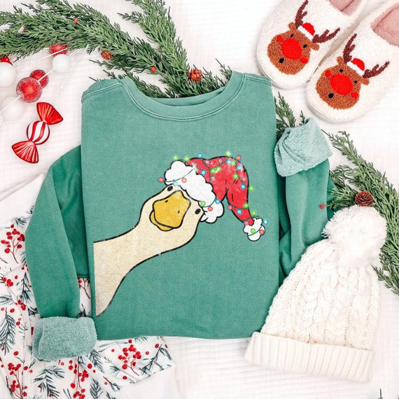 Christmas Goose DTF Transfer | Funny Santa Hat Holiday Design, festive goose wearing a light-up Santa hat printed on a teal sweatshirt surrounded by Christmas greenery, reindeer slippers, and cozy winter accessories