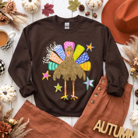 Thankful Turkey DTF Transfer | Colorful Fall Thanksgiving Design, colorful turkey with patterned feathers, stars, and “thankful” lettering displayed on a brown sweatshirt in an autumn-themed flat lay