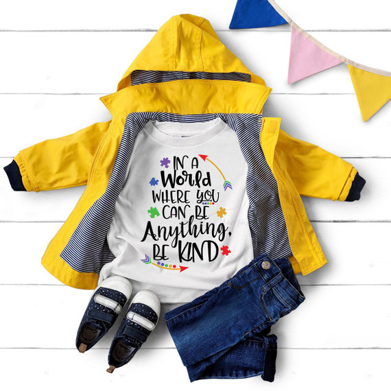 Product, In a world where you can be anything be kind Puzzle Autism Awareness Sublimation Transfer T-shirt with colorful puzzle pieces and inspirational quote, displayed with a yellow jacket, jeans, and sneakers on a white background.