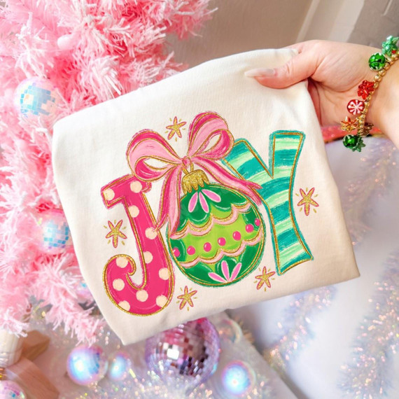 JOY Christmas DTF Transfer | Preppy Pink & Green Ornament Design, colorful pink and green JOY lettering with a decorated Christmas ornament and bow printed on a white shirt beside a pink Christmas tree.