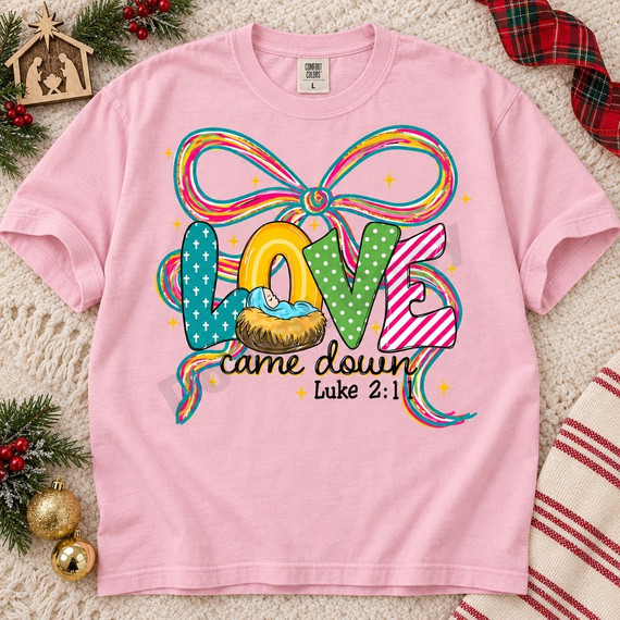 LOVE came down Christmas DTF transfer with patterned bubble letters, swaddled baby in a manger, multicolor ribbon bow, and Luke 2:11 scripture Shown on a Blossom t-shirt.