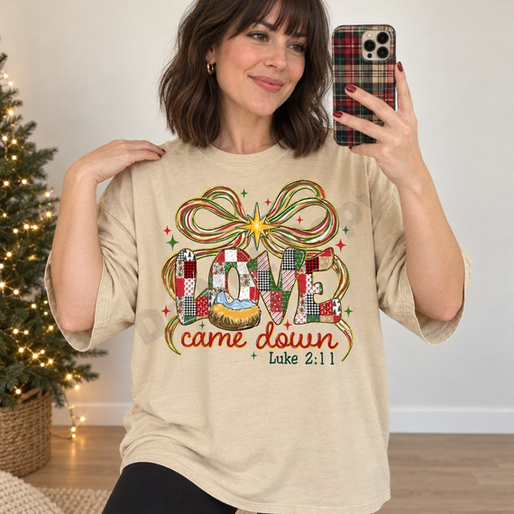 Love Came Down nativity DTF transfer with patchwork LOVE lettering, baby Jesus in a manger, holiday bow with glowing star, and Luke 2:11 Bible verse Shown on a Sandstone t-shirt.