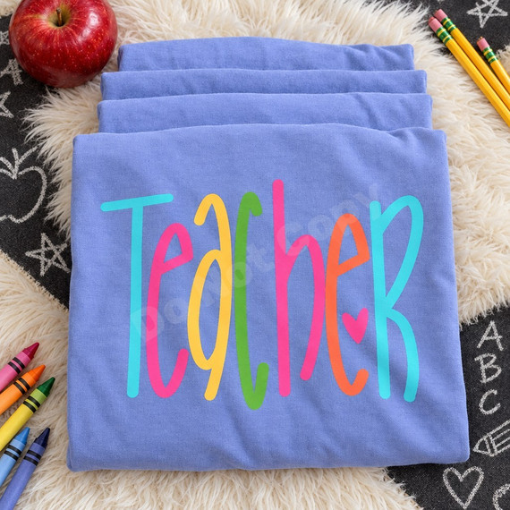 Colorful hand-lettered Teacher DTF transfer in rainbow brush script with a pink heart accent Shown on a Periwinkle t-shirt.