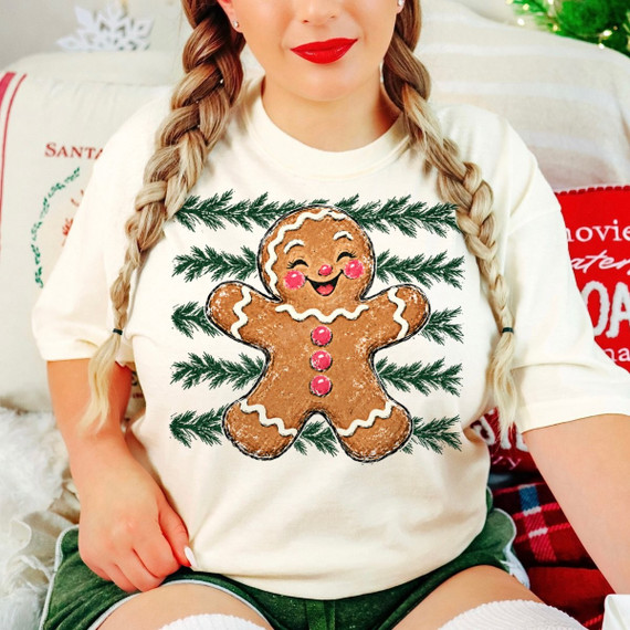 Smiling gingerbread man DTF transfer with icing details and pine branch sprigs on a cream t-shirt