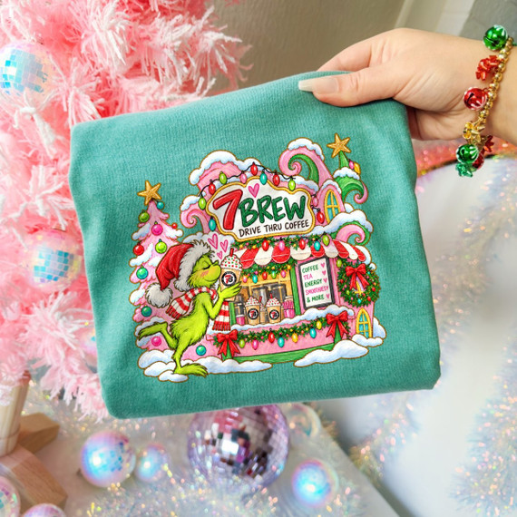 Green holiday grinch character sipping a drink beside a pink drive-thru coffee shop DTF transfer with festive Christmas lights and wreaths