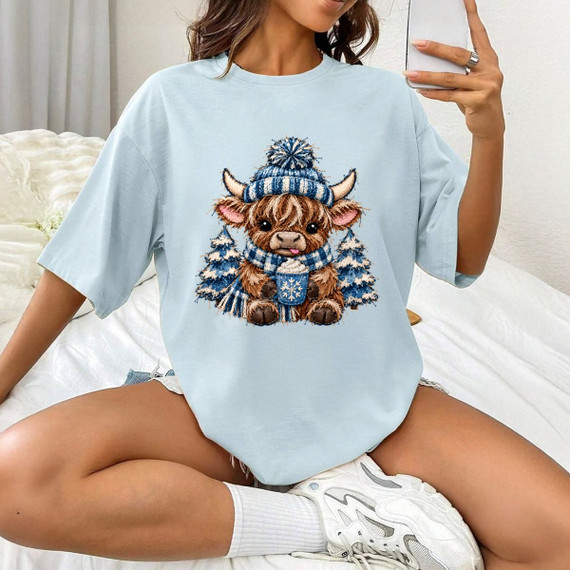 Winter highland cow DTF transfer wearing a striped beanie and plaid scarf, holding hot cocoa with snowy pine trees, embroidery-style design