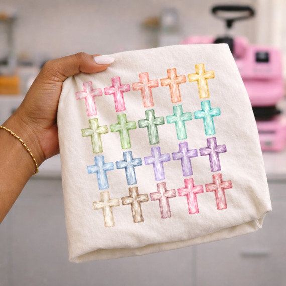 DTF transfer featuring a grid of 20 hand-sketched crosses in rainbow watercolor colors on a cream shirt