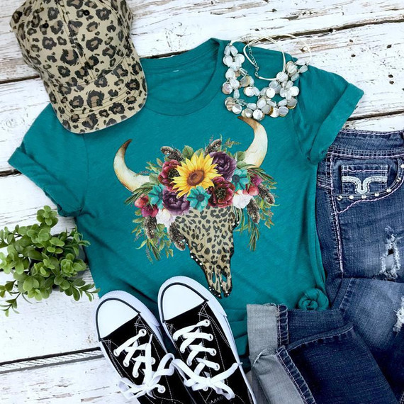 Leopard Bull Skull with Floral Headpiece Screen Print Transfer, casual outfit with teal t-shirt, leopard print cap, distressed jeans, black sneakers, and jewelry, on white wooden surface.
