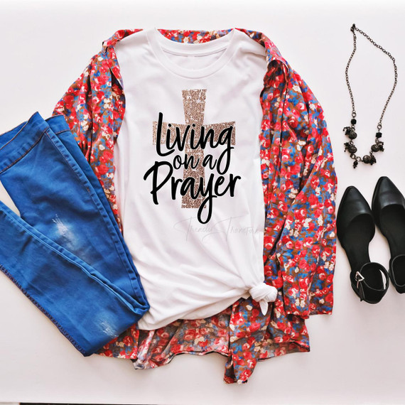 Living On A Prayer with gold foil effect Cross Sublimation Transfer, product display featuring white t-shirt with black and gold graphic design, paired with blue jeans, black shoes, jewelry, and a floral shirt.