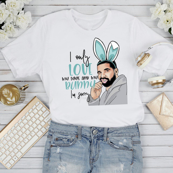 Product: I Only Love My Eggs and My Bunny, I’m Sorry Drake Easter Parody Sublimation Transfer; image features a white t-shirt with a cartoon illustration of a man with a beard wearing bunny ears, with text saying 'I only love my eggs and my bunny, I’m sorry Drake', laid out on a white wooden surface with a bouquet of white flowers, a gold-rimmed coffee mug, a beige keyboard, white headphones, and a gold wallet nearby.