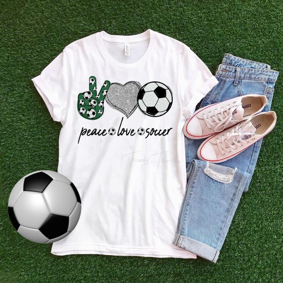 Peace Love Soccer heart glitter Sublimation Transfer, white t-shirt with soccer-themed graphic design on green artificial grass with pair of ripped jeans, pink shoes, and a soccer ball.