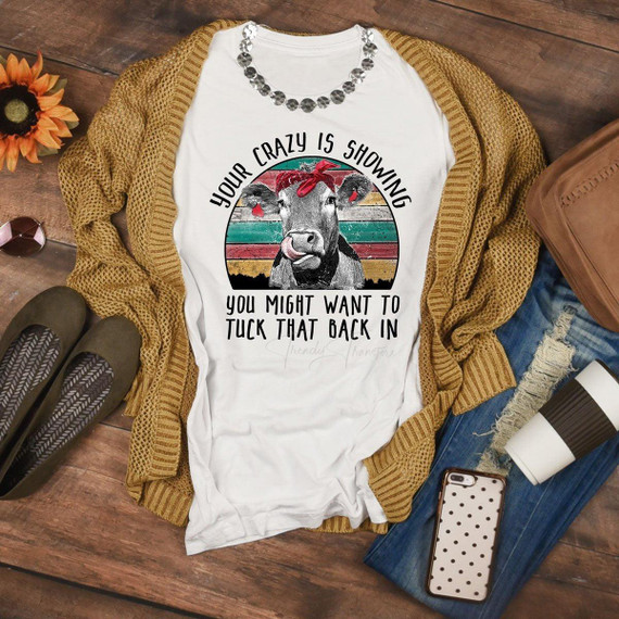 Your Crazy is Showing Cow Sublimation Transfer, humorous cow graphic with text on a white T-shirt, surrounded by fashion accessories and casual items on a rustic wooden surface.