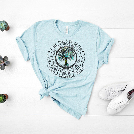 I See Trees of Green Screen Print Heat Transfer, a light blue T-shirt with a circular tree graphic and poetic text about trees and blooming, laid flat on a white wooden surface with small potted succulents and white sneakers nearby.