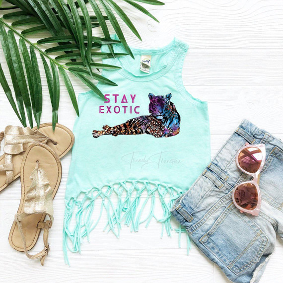 Stay Exotic FINAL STOCK - YOUTH Screen Print Heat Transfer, youth tank top with a colorful tiger and lion graphic and fringed hem, surrounded by casual summer accessories including sandals, sunglasses, and denim shorts on a white wooden surface.