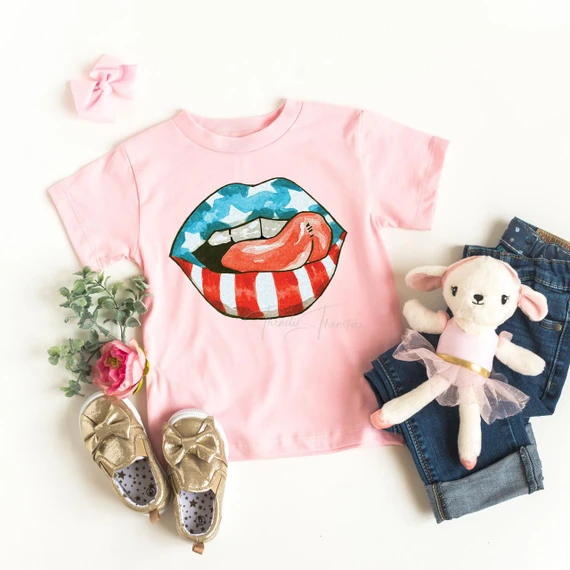 Patriotic Lips and Tongue Youth Screen Print Heat Transfer, pink T-shirt with a graphic of lips with an American flag pattern and tongue, surrounded by children's accessories including gold shoes, jeans, a plush doll, a pink bow, and flowers.