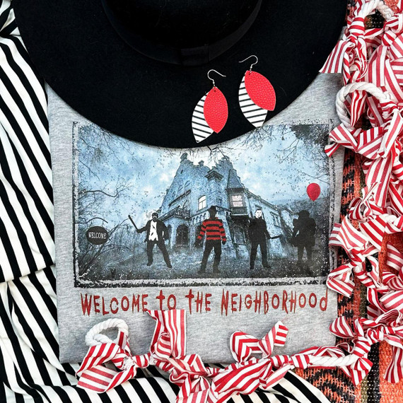 Welcome To The Neighborhood Screen Print Transfer, a design featuring a spooky haunted house with four figures, balloons, and the phrase 'Welcome to the Neighborhood' in red text, laid on a gray fabric surrounded by striped ribbons and accessories.