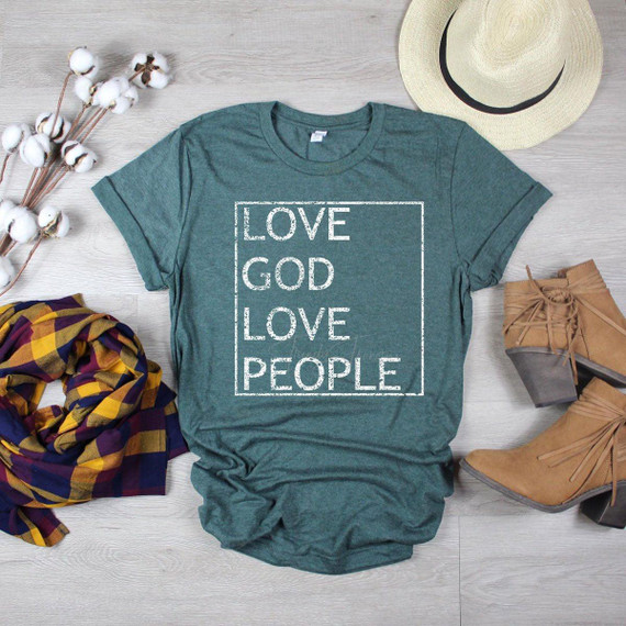 Love God Love People Distressed Screen Print Transfer T-shirt, casual teal shirt with white distressed text, surrounded by a straw hat, ankle boots, cotton branch, and a plaid scarf on a light wooden surface.