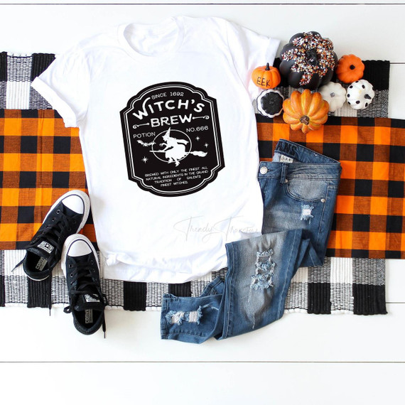 Witch's Brew Potion Sublimation Transfer, Halloween-themed black and white graphic design on a white T-shirt, displayed alongside pumpkins, a pair of black and white sneakers, and distressed denim jeans on a checkered orange and black tablecloth.