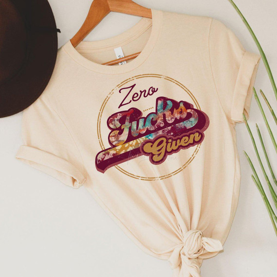 Zero F*cks Given Screen Print Heat Transfer T-shirt with colorful, bold graphic design on a beige T-shirt, knotted at the bottom, laid flat on a white surface next to a black hat and green plant leaves.