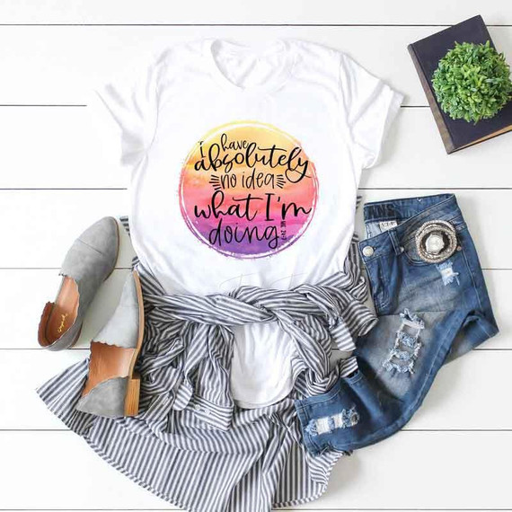 I Have Absolutely No Idea Sublimation Transfer, white T-shirt with colorful round design and text print, laid on a striped fabric, with ripped jeans, gray shoes, and a small potted plant surrounding it.