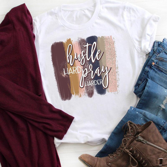 Hustle Hard Pray Harder Brushstrokes Sublimation Transfer, graphic t-shirt with motivational quote and colorful brushstroke background, paired with jeans and brown boots on a white surface.