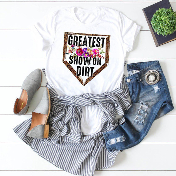 Greatest Show On Dirt Leopard Sublimation Transfer, white t-shirt with a leopard print border and colorful floral design, laid out with denim jeans, gray shoes, and a striped shirt on a white wooden surface with a black book and a green plant.