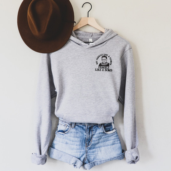 Not Fragile Like a Flower RBG POCKET/Mask Screen Print Transfer, front view of a gray hoodie with black text and graphic design on the chest, paired with denim shorts and a brown hat hanging on a hanger.