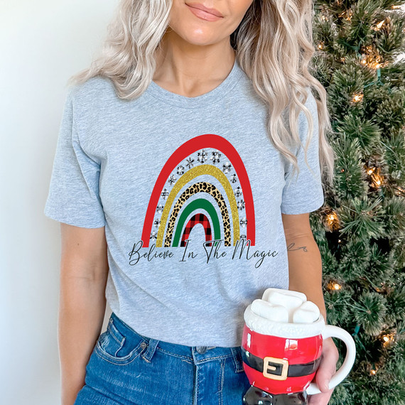 Believe In The Magic Screen Print Heat Transfer, a colorful rainbow design with various patterns and the phrase 'Believe In The Magic' on a light grey t-shirt worn by a woman.