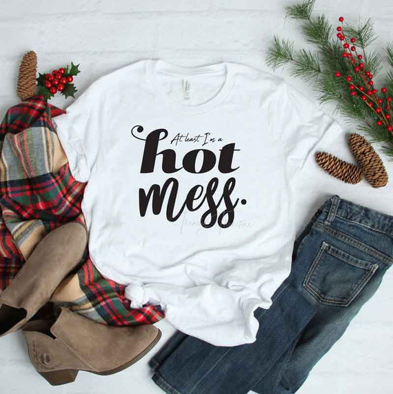 At Least I'm a Hot Mess sublimation transfer, white t-shirt with black text, holiday-themed flat lay with plaid blanket, boots, pine branches, and pinecones
