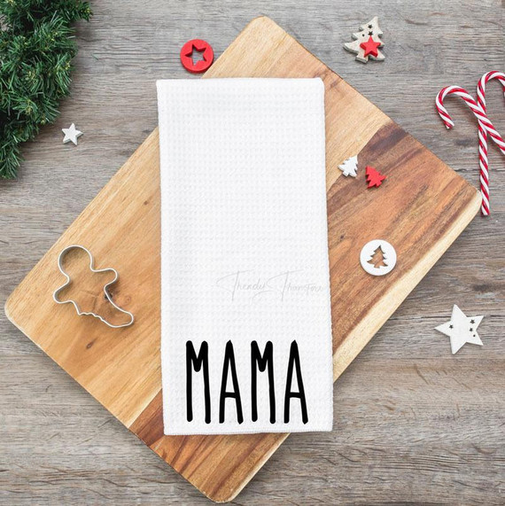Mama Hat/Tea Towel Screen Print Heat Transfer, white tea towel with black 'MAMA' text, surrounded by holiday decorations on wooden surface.