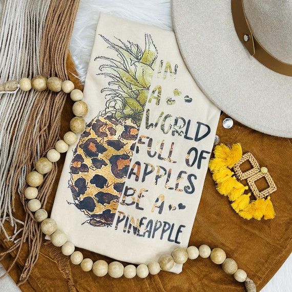 In a World Full Of Apples Be a Pineapple Screen Print Heat Transfer, a clothing embellishment featuring a pineapple graphic with motivational text, displayed alongside fashion accessories on a wooden surface.