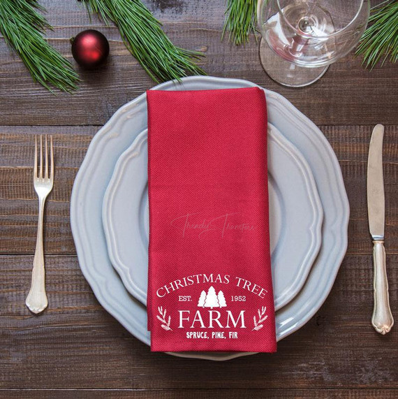 Product Christmas Tree Farm Tea Towel Screen Print Heat Transfer, red tea towel with Christmas tree farm design on a place setting with silverware, glass, and holiday decor.