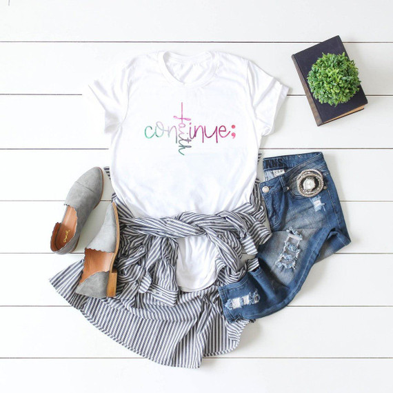 Continue Suicide Awareness Sublimation Transfer, white T-shirt with colorful text design, denim shorts, gray slip-on shoes, a striped shirt tied around the waist, and a small plant on a white wooden surface.