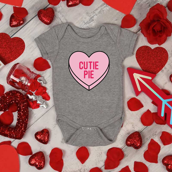 Cutie Pie Candy Heart INFANT Screen Print Heat Transfer on a gray baby onesie surrounded by red decorative hearts and Valentine's Day-themed items.
