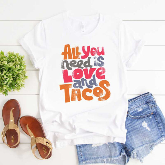 All You Need Is Love And Tacos Sublimation Transfer T-shirt with colorful text design, laid flat with denim shorts, sandals, and a small green plant on white wooden background.