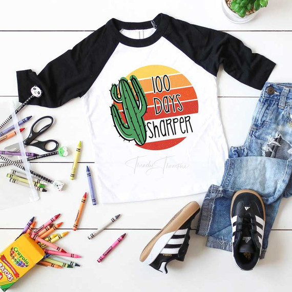 100 Days Sharper Sublimation Transfer, a graphic T-shirt with a cactus and sunset design, laid out with Crayola crayons, scissors, and casual denim and sneakers on a white wooden surface.