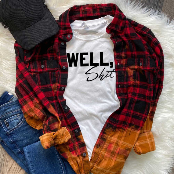 Well, Shit Sublimation Transfer, white t-shirt with black and cursive text design, styled with red and black plaid shirt, jeans, and a black cap on a cozy white rug.