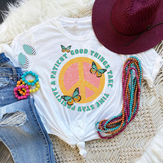 Good Things Take Time Sublimation Transfer, colorful peace sign t-shirt with butterfly graphics, paired with denim shorts, vibrant jewelry, and a wide-brimmed red hat, all laid out on a textured surface.