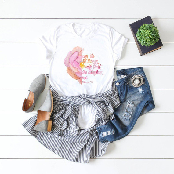 I Can Do All Things Through Christ Rose Sublimation Transfer, T-shirt with inspirational Bible verse and pink rose graphic, paired with striped shirt, distressed jeans, grey flats, black book with green plant on top, on white wooden background.