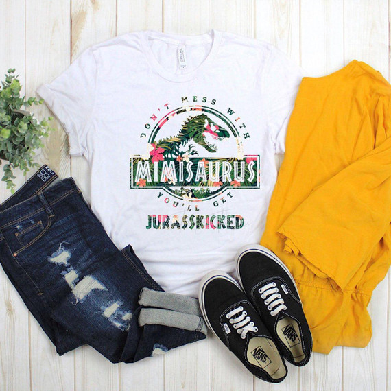 Don't Mess With Mimisaurus You'll Get Jurasskicked Tropical Sublimation Transfer T-shirt, featuring a colorful dinosaur graphic with floral accents and bold text, laid flat alongside yellow pants, black Vans sneakers, a pair of distressed jeans, and a green plant, on a light wooden surface.