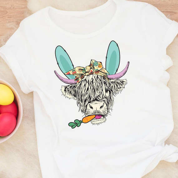 Shaggy Easter Cow Sublimation Transfer, a graphic design of a cow with shaggy fur, rainbow-colored ears and antlers, a bow on its head, and holding a carrot in its mouth on a white t-shirt.