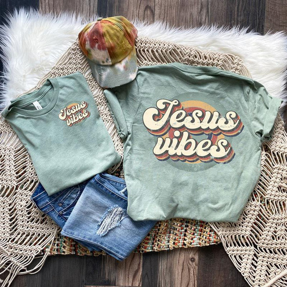Jesus Vibes POCKET Screen Print Heat Transfer, showcasing a casual outfit with a large graphic t-shirt, a smaller matching t-shirt, distressed denim shorts, and a tie-dye cap laid out on a woven blanket.